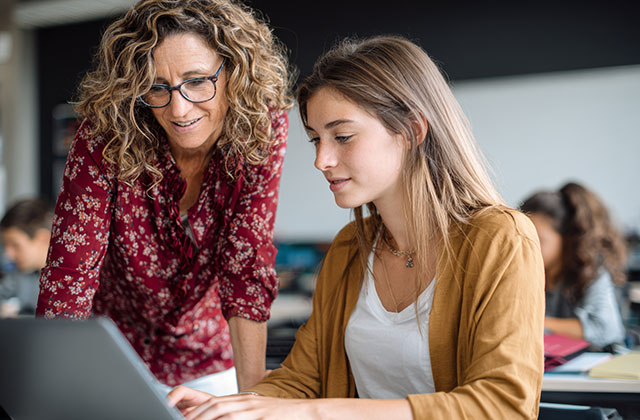 a retired teacher working as a substitute helps a student with a task on a computer