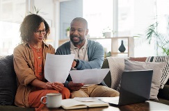 a couple sitting on a couch smiling while looking at paperwork with a laptop computer