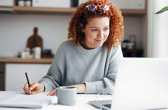 a woman using a laptop and taking notes in a notebook