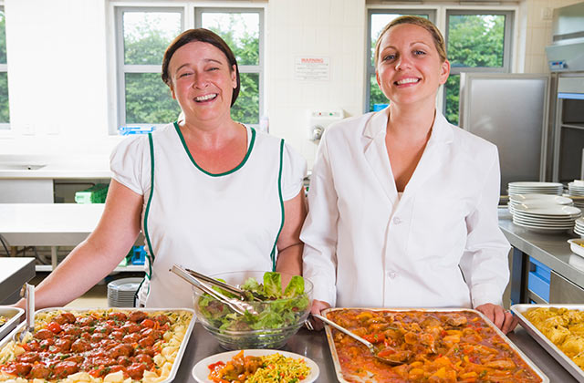 two school cafeteria workers posing for the camera in the kitchen with platters of food