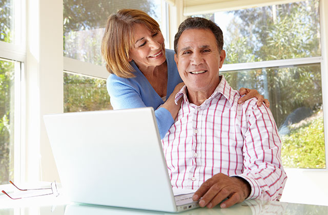 middle-aged couple sitting in a brightly lit kitchen with a laptop computer smiling