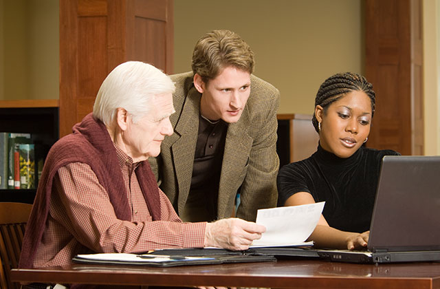 older gentleman and adult grandson looking at paperwork as a benefit counselor types on a computer