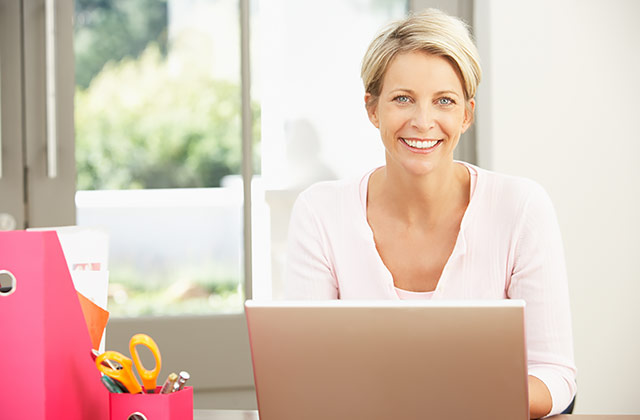 woman sitting at a craft table while typing on a laptop computer and smiling at the camera