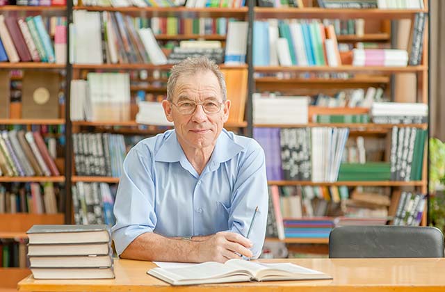 teacher sitting at a desk in a library with a stack of books to his right and an open book in front of him